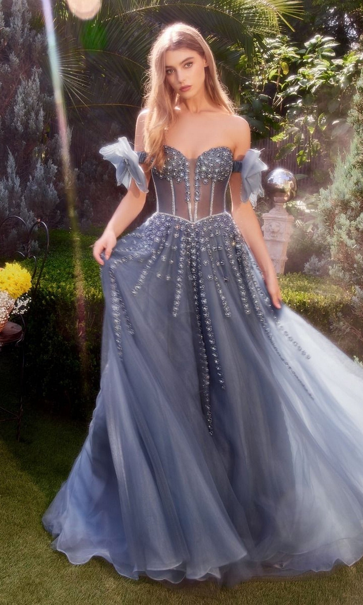A woman in a strapless Andrea and Leo A1338 long prom dress stands on grass in a sunlit garden, gracefully holding the sides of her beaded evening gown.